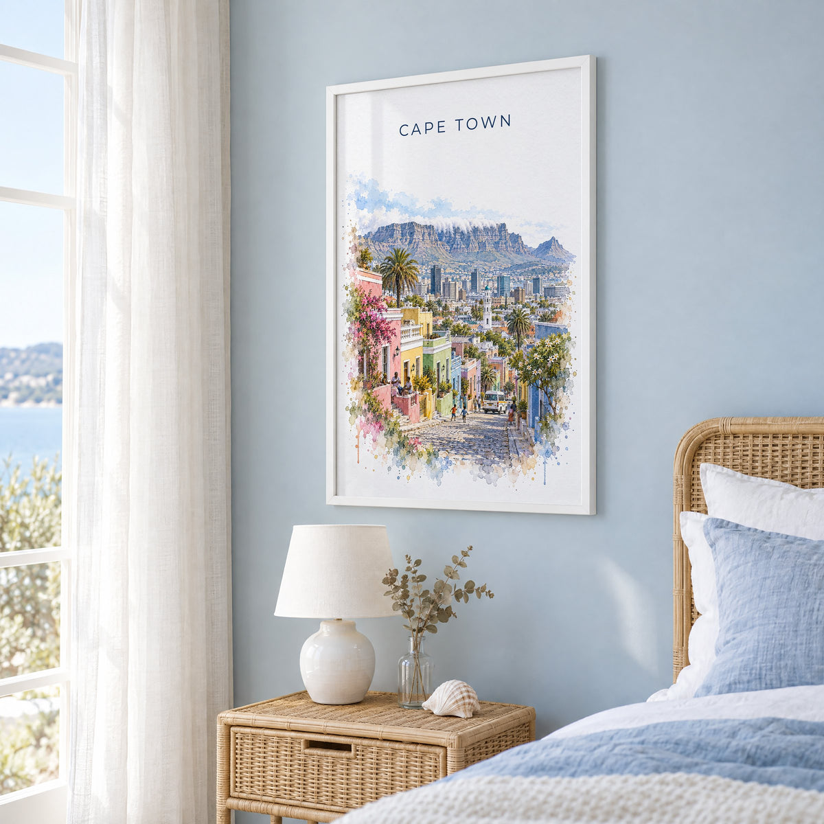 Cape Town watercolor print in a white frame on a pale blue wall beside a rattan bed in a coastal bedroom