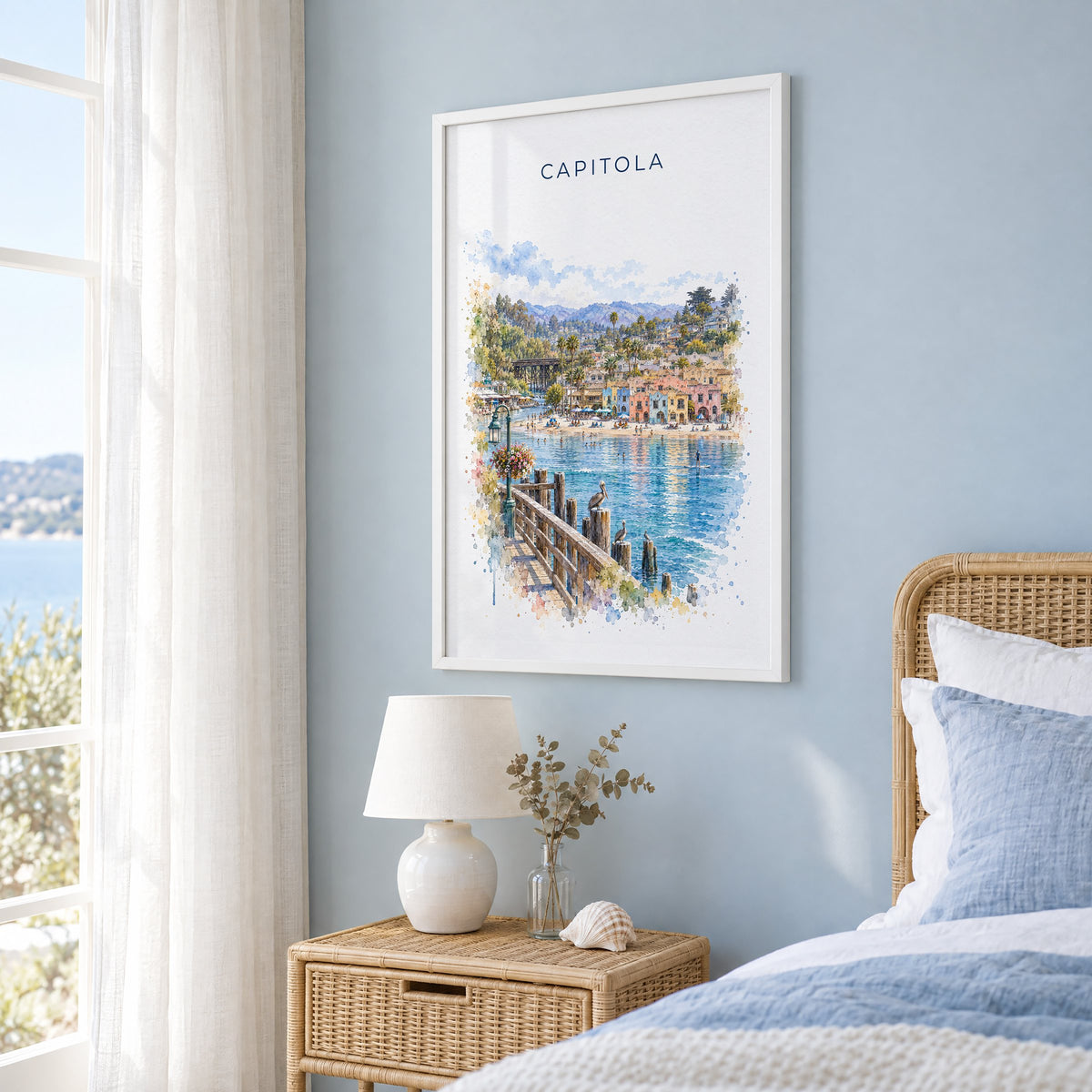 Capitola, California watercolor print in a white frame on a pale blue wall beside a rattan bed in a coastal bedroom