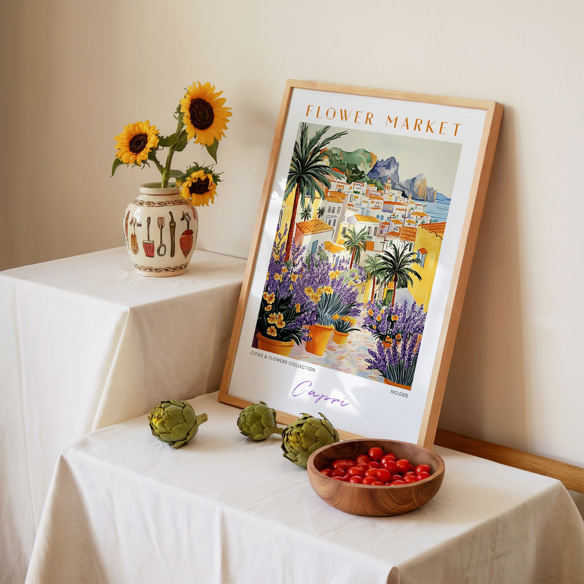 Capri flower market print in an oak frame leaning on a linen-draped table beside a vase of sunflowers