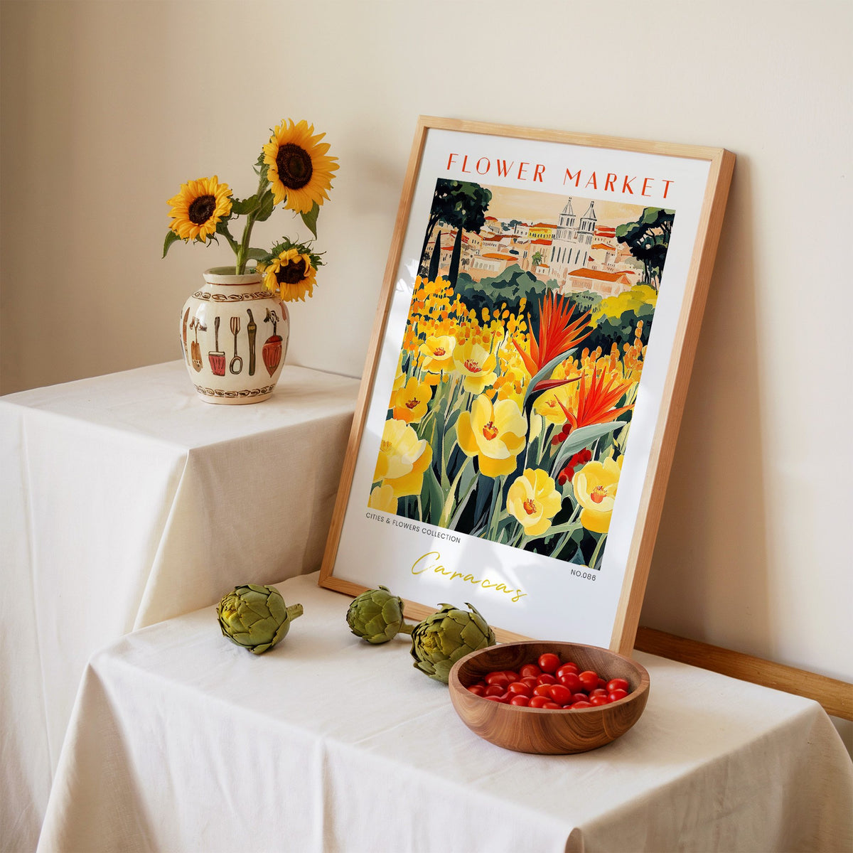 Caracas flower market print in an oak frame leaning on a linen-draped table beside a vase of sunflowers