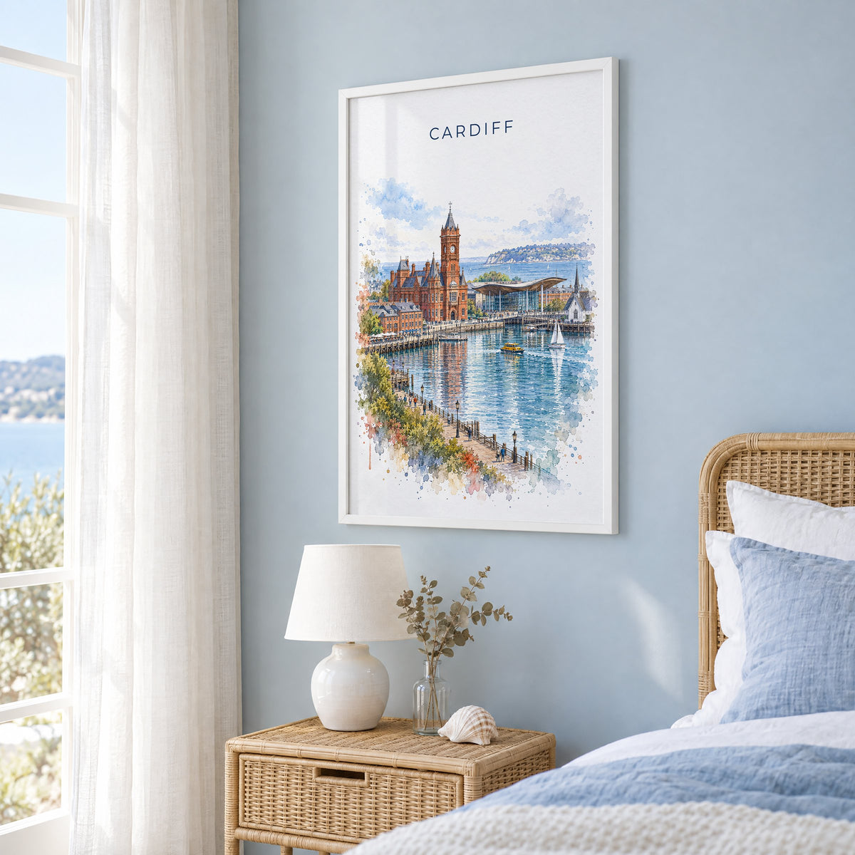 Cardiff watercolor print in a white frame on a pale blue wall beside a rattan bed in a coastal bedroom