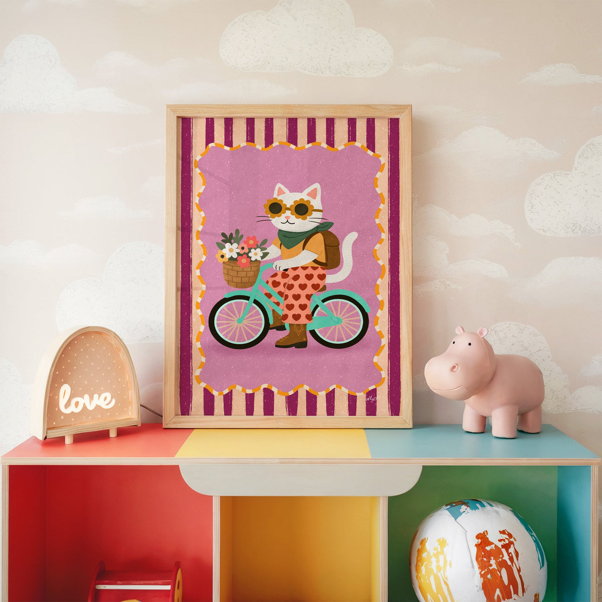 White cat riding a mint bicycle nursery print in an oak frame on a rainbow toy shelf against cloud wallpaper