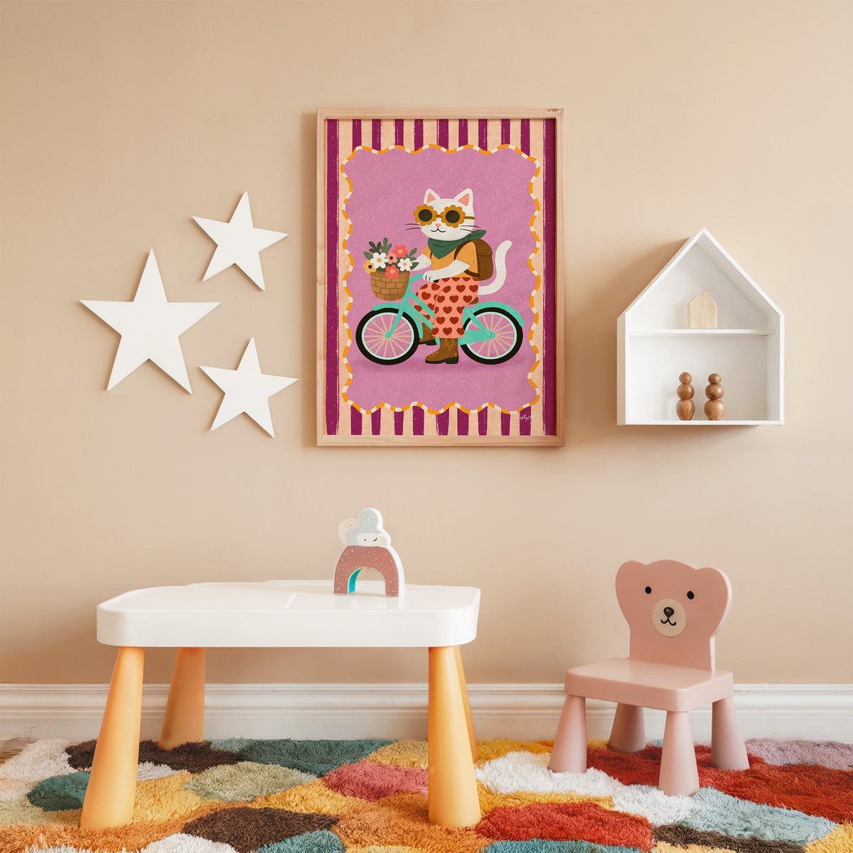 White cat riding a mint bicycle nursery print in an oak frame on a peach wall above a kids table with wall stars
