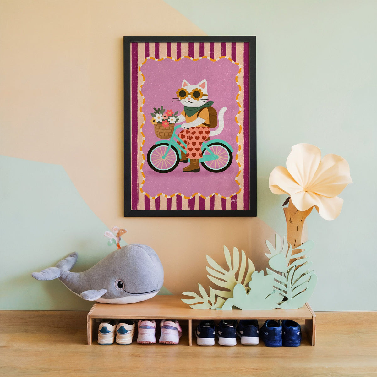 White cat riding a mint bicycle nursery print in a black frame on a mint and peach wall above a whale plush