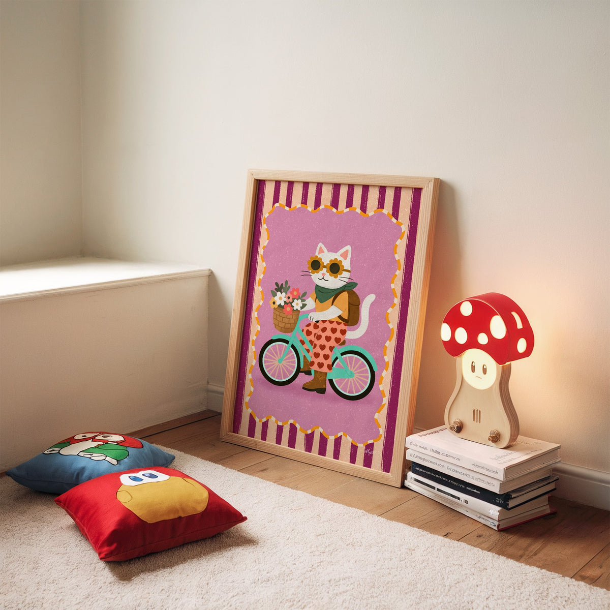White cat riding a mint bicycle nursery print in an oak frame leaning on a rug beside a mushroom lamp and floor cushions