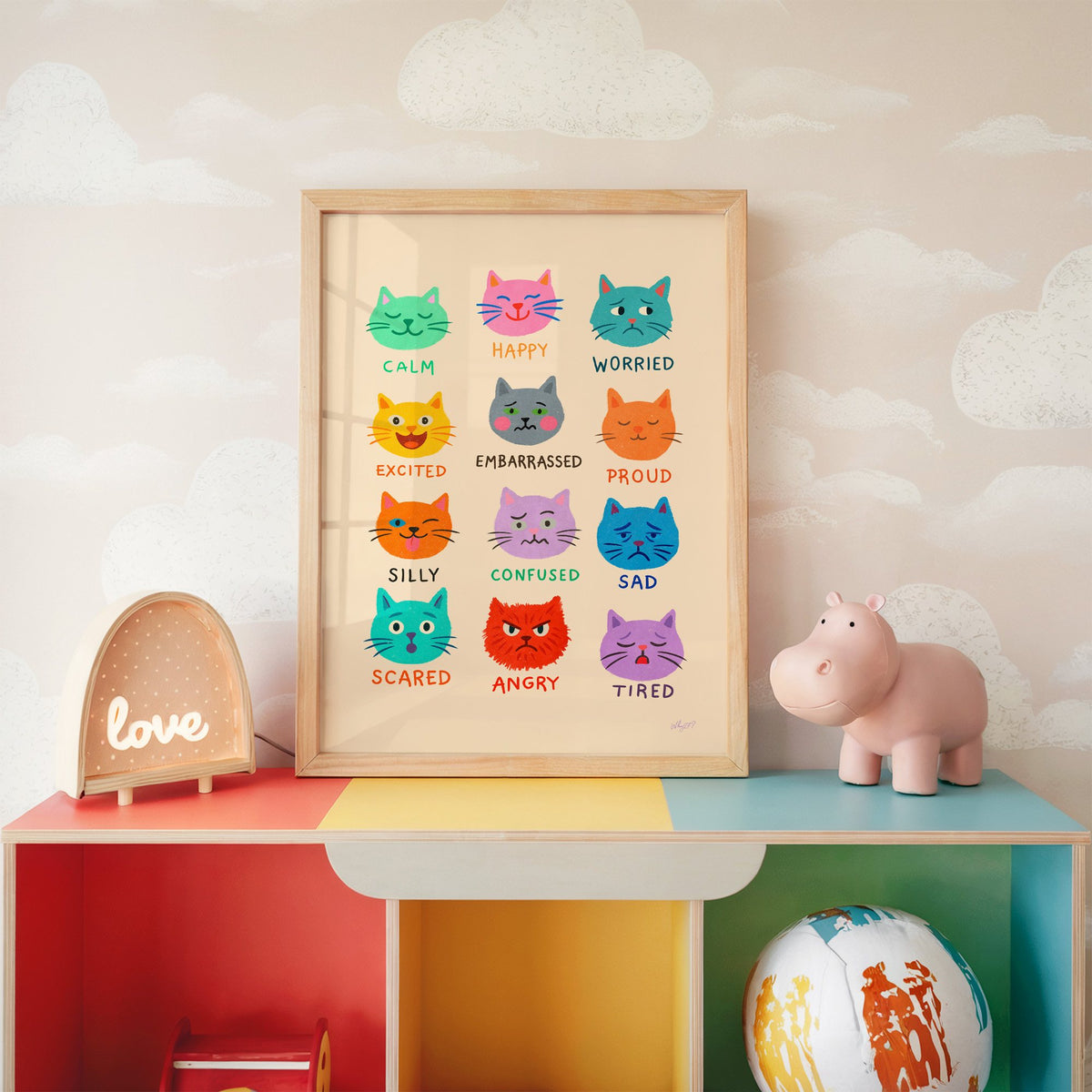 Colourful cat feelings chart nursery print in an oak frame on a rainbow toy shelf against cloud wallpaper