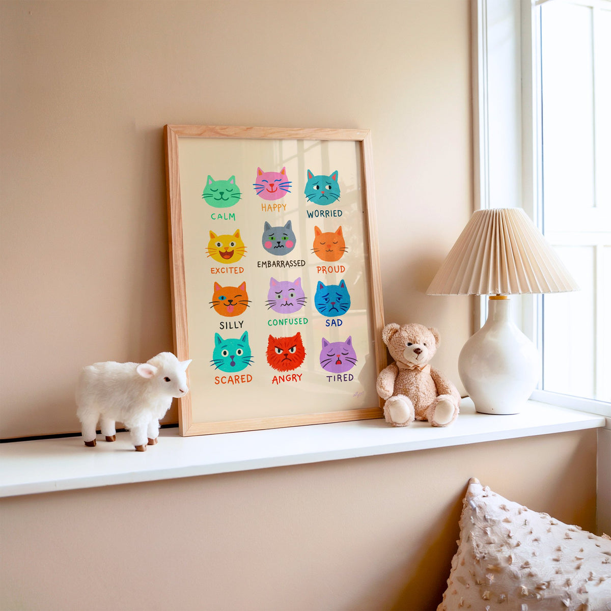 Colourful cat feelings chart nursery print in an oak frame on a window ledge with a toy lamb and teddy