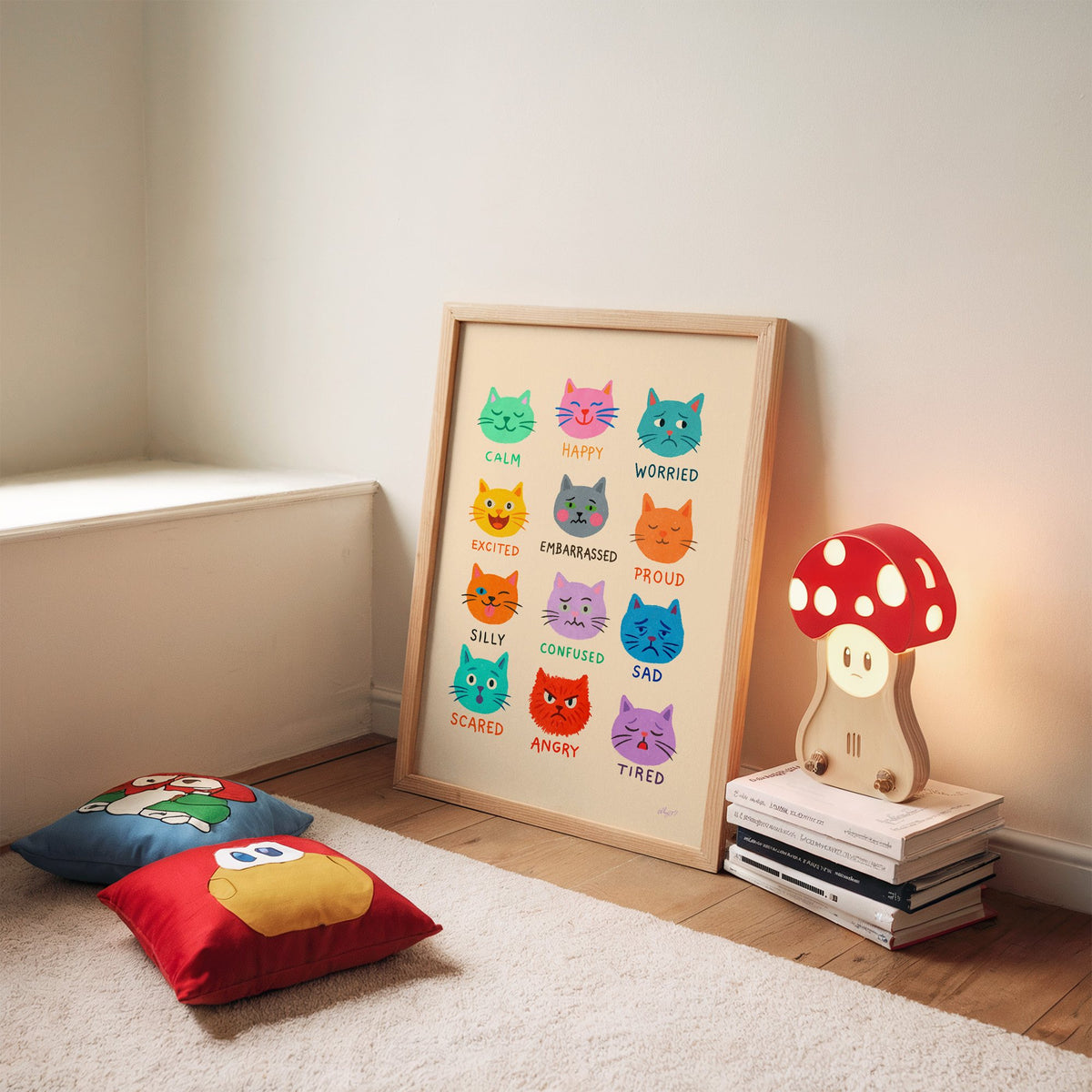 Colourful cat feelings chart nursery print in an oak frame leaning on a rug beside a mushroom lamp and floor cushions