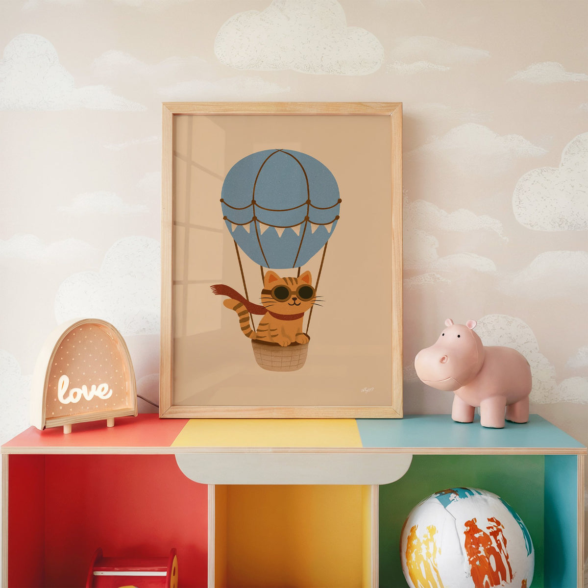 Cat in blue hot air balloon nursery print in an oak frame on a rainbow toy shelf against cloud wallpaper