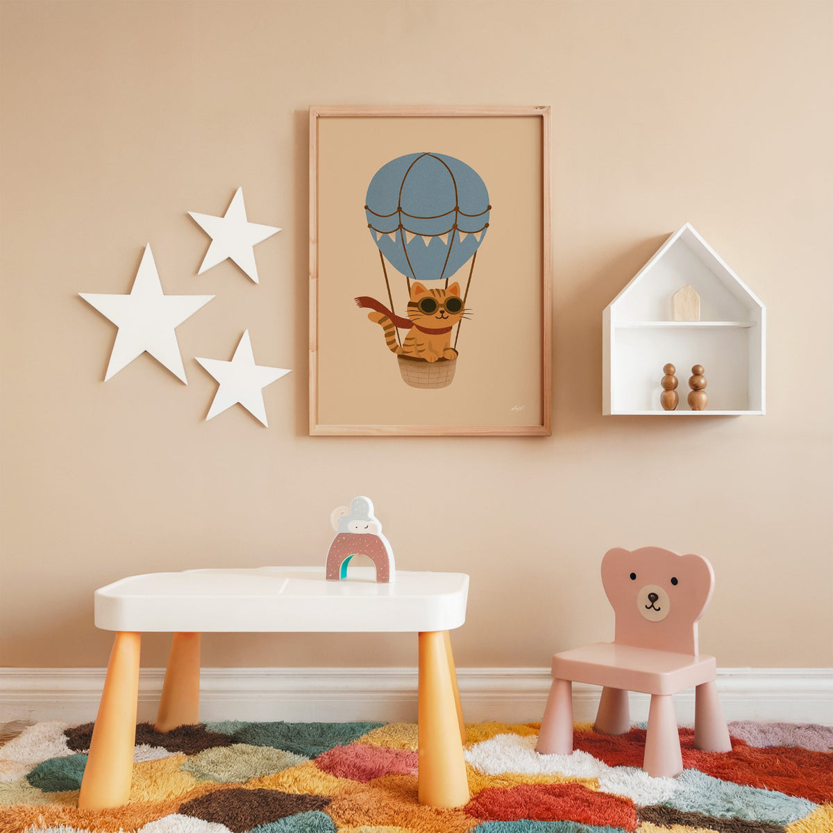 Cat in blue hot air balloon nursery print in an oak frame on a peach wall above a kids table with wall stars