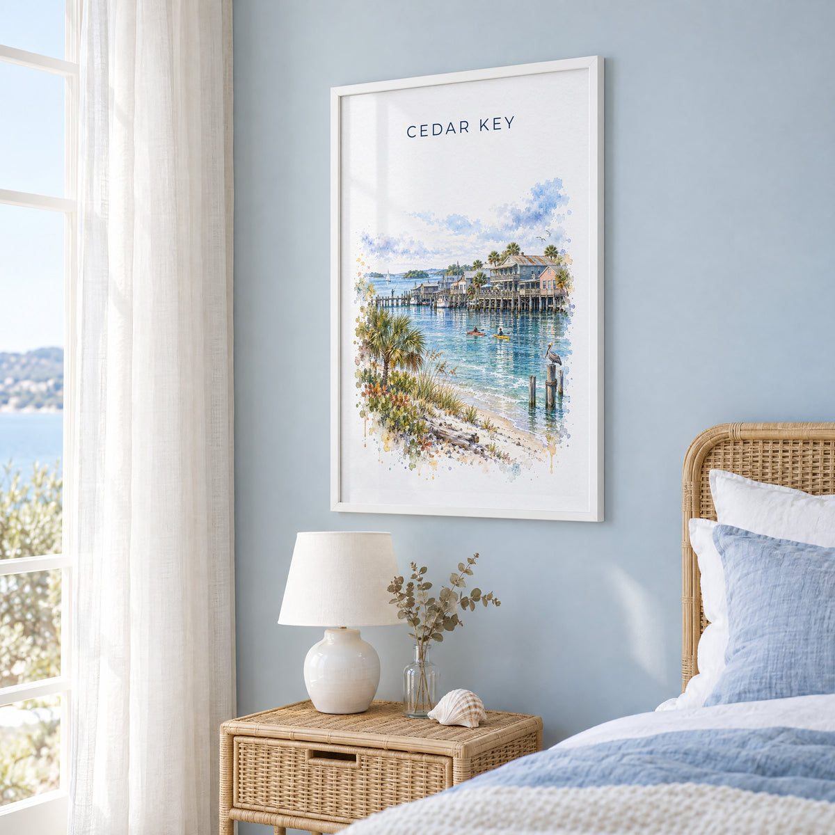 Cedar Key, Florida watercolor print in a white frame on a pale blue wall beside a rattan bed in a coastal bedroom