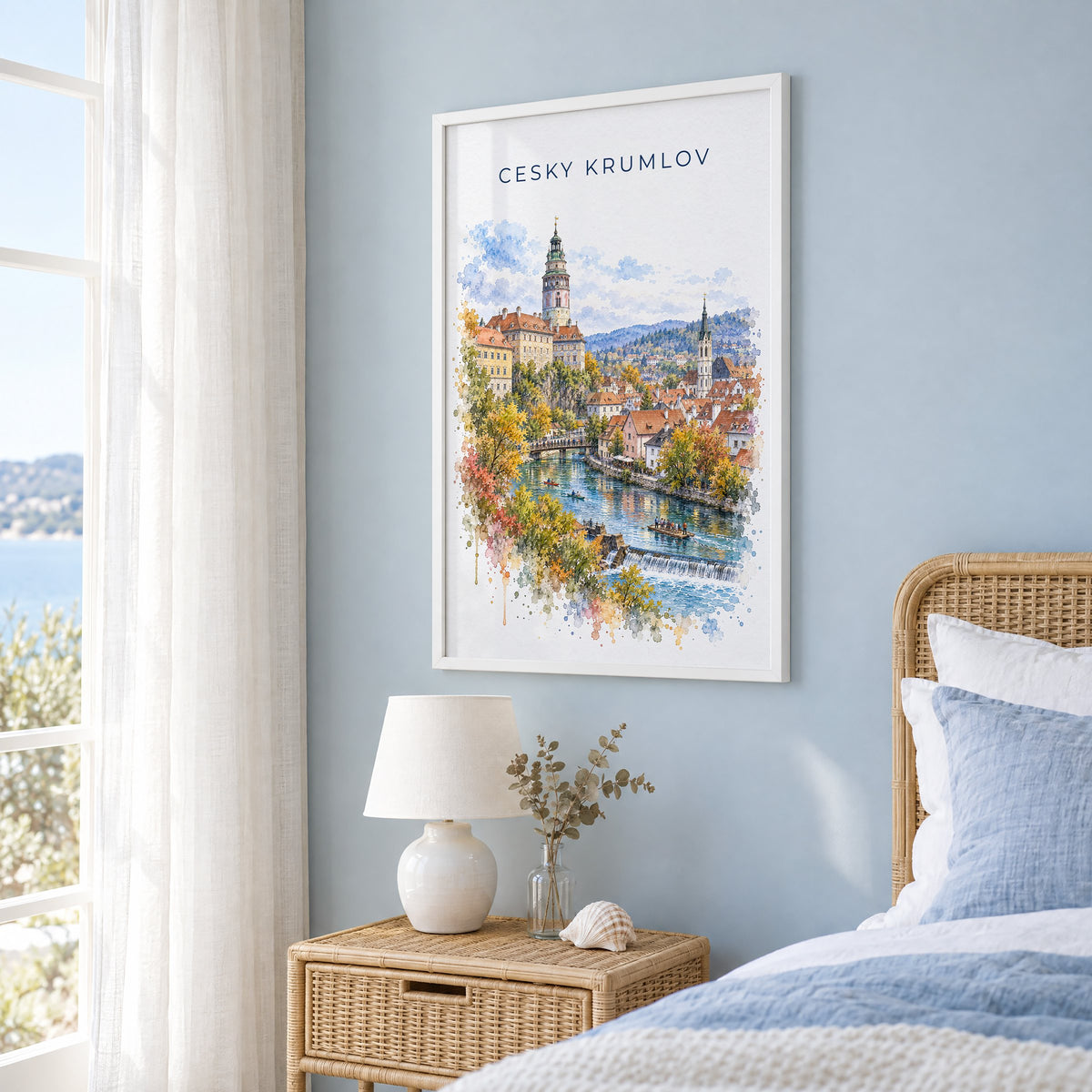 Cesky Krumlov watercolor print in a white frame on a pale blue wall beside a rattan bed in a coastal bedroom