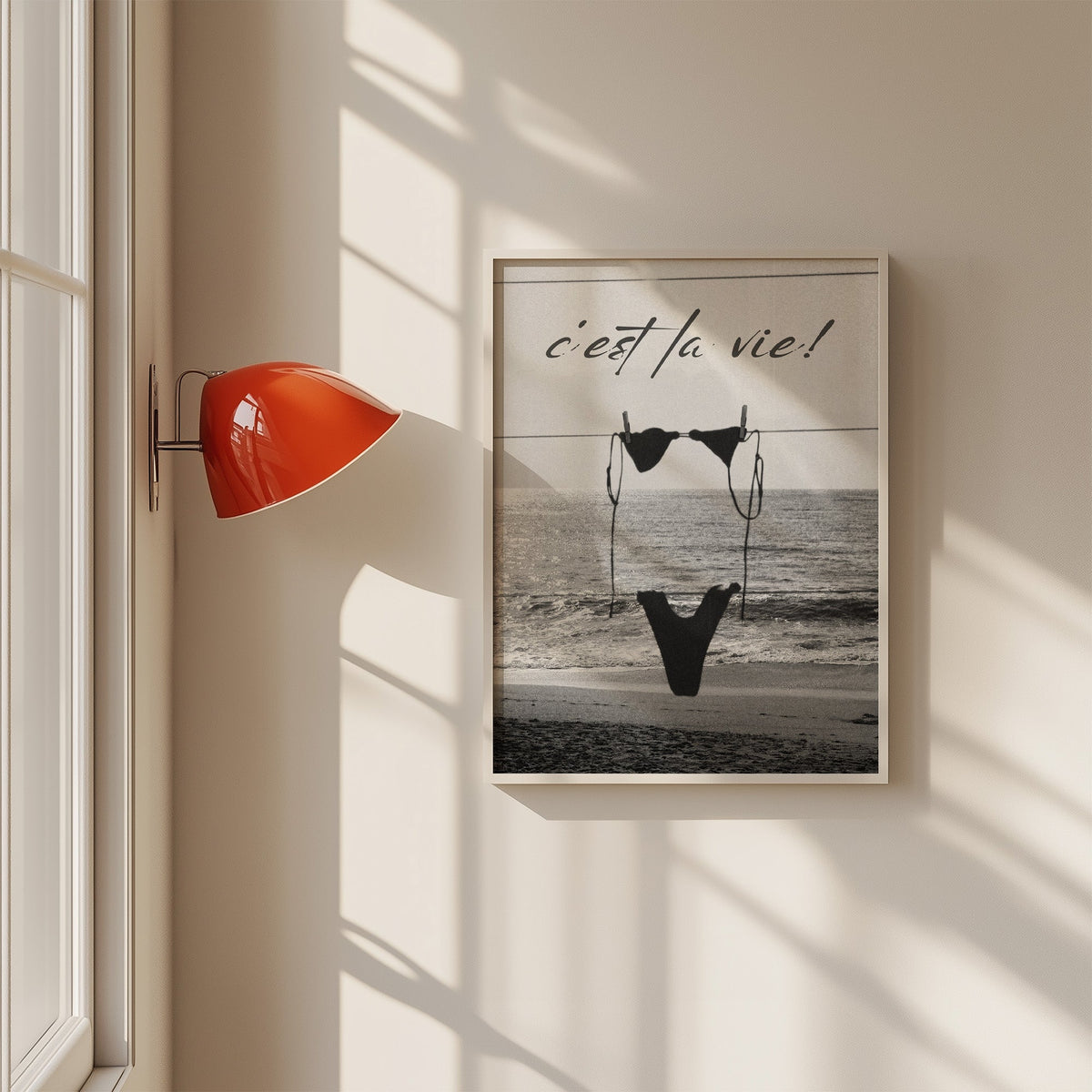 Black bikini on a clothesline over the sea print in a white frame on a sunlit wall beside a red sconce