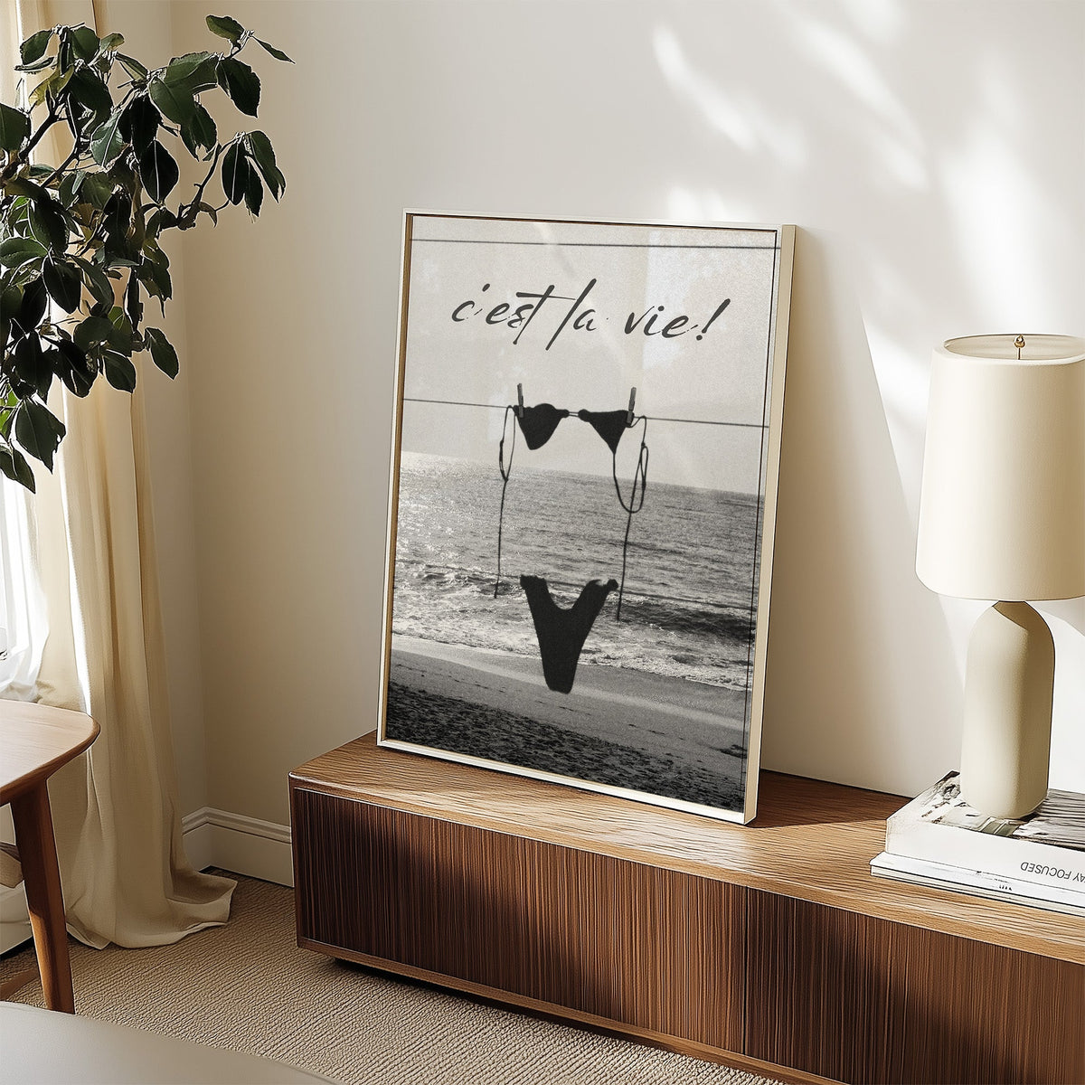 Black bikini on a clothesline over the sea print in a white frame on a walnut sideboard beside a ceramic lamp