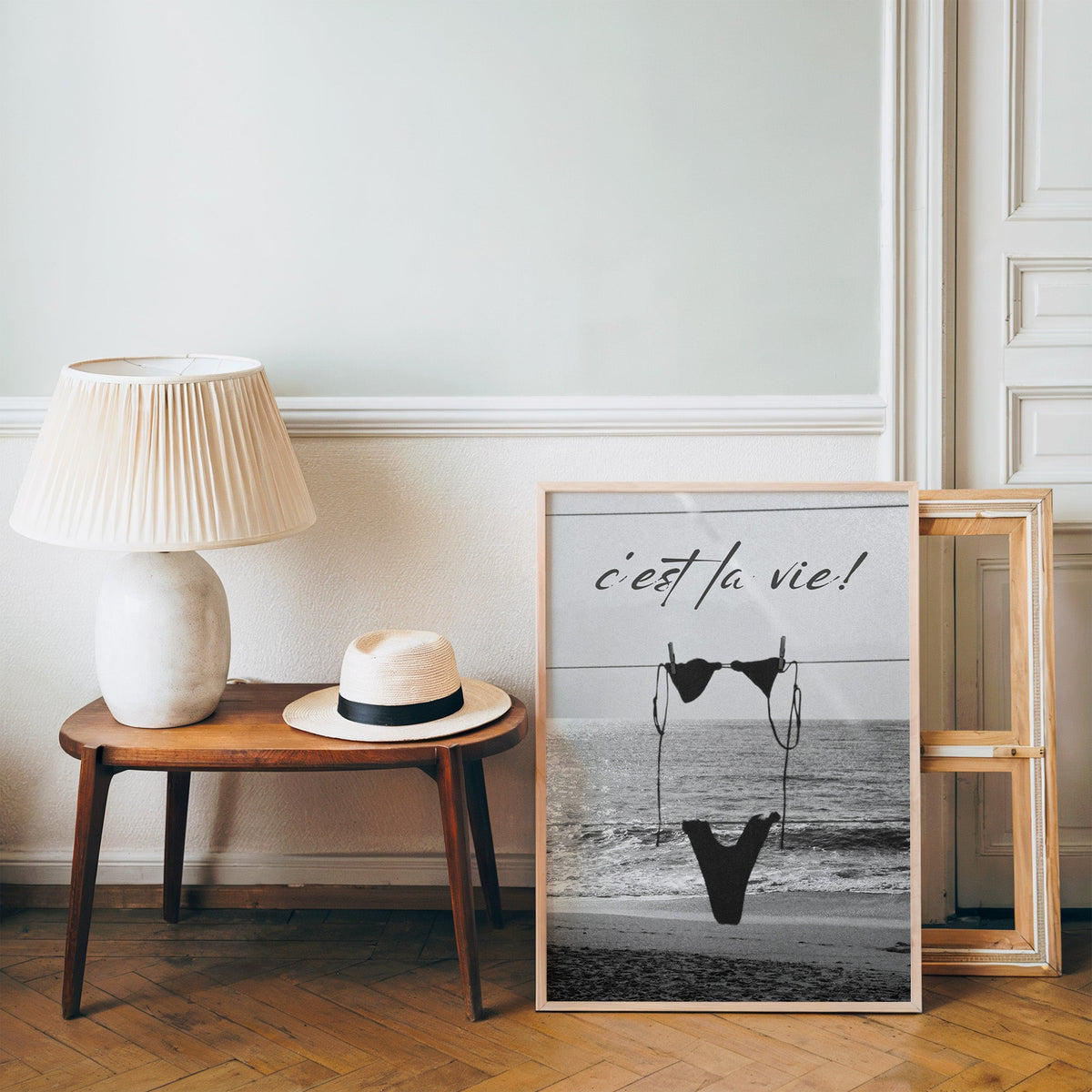 Black bikini on a clothesline over the sea print in a pine frame on a wood floor beside a lamp and straw hat