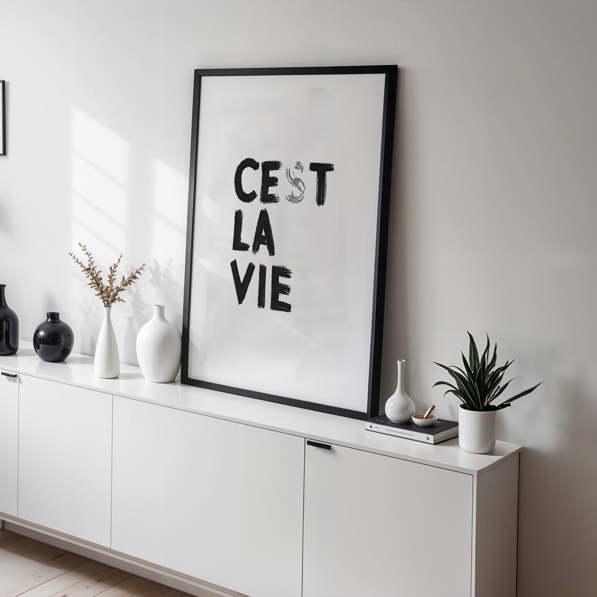 Cest la vie brush lettering on white print in a black frame on a white sideboard with black and white vases