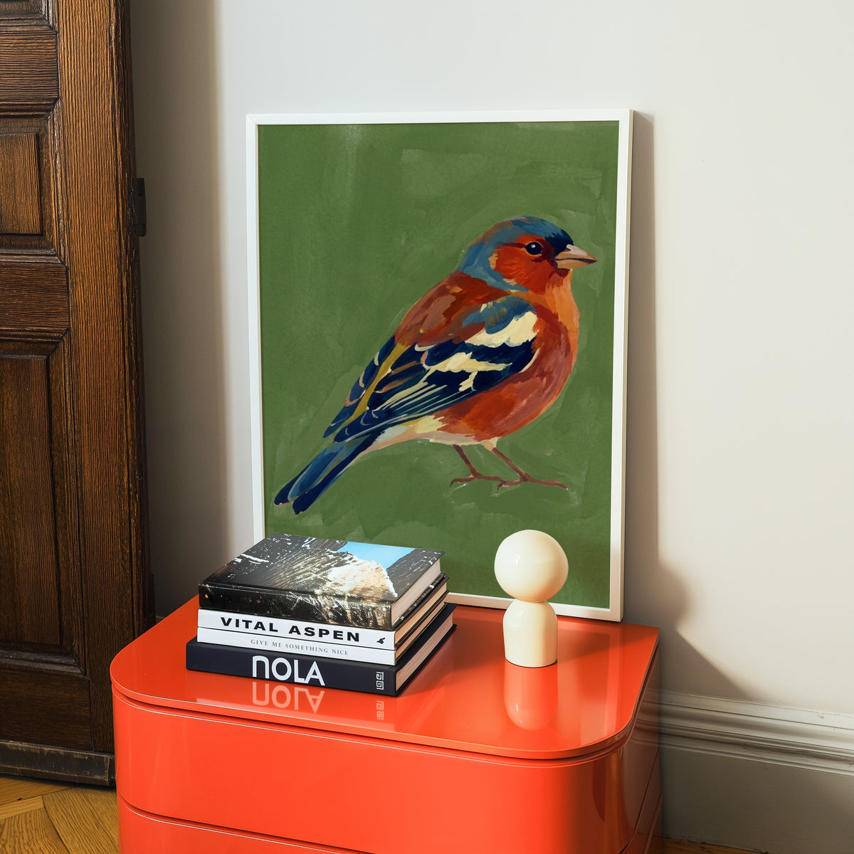 Painted songbird print in a white frame on a glossy orange cabinet with books