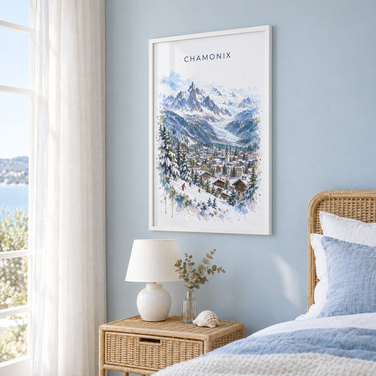 Chamonix watercolor print in a white frame on a pale blue wall beside a rattan bed in a coastal bedroom