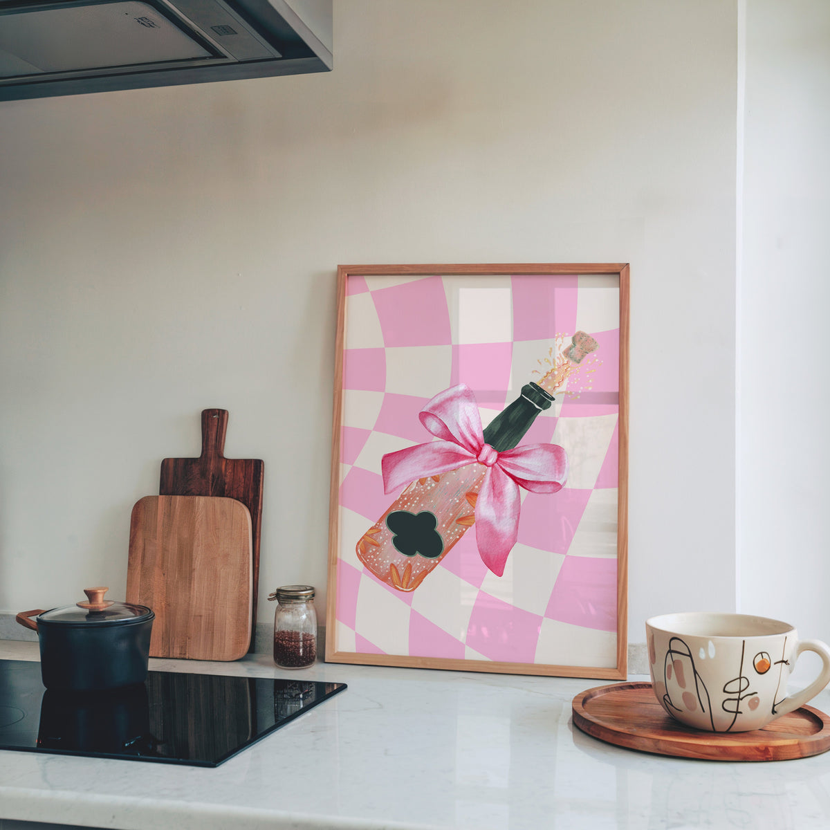 Champagne bottle with a pink bow print in a pine frame leaning on a marble kitchen counter beside chopping boards