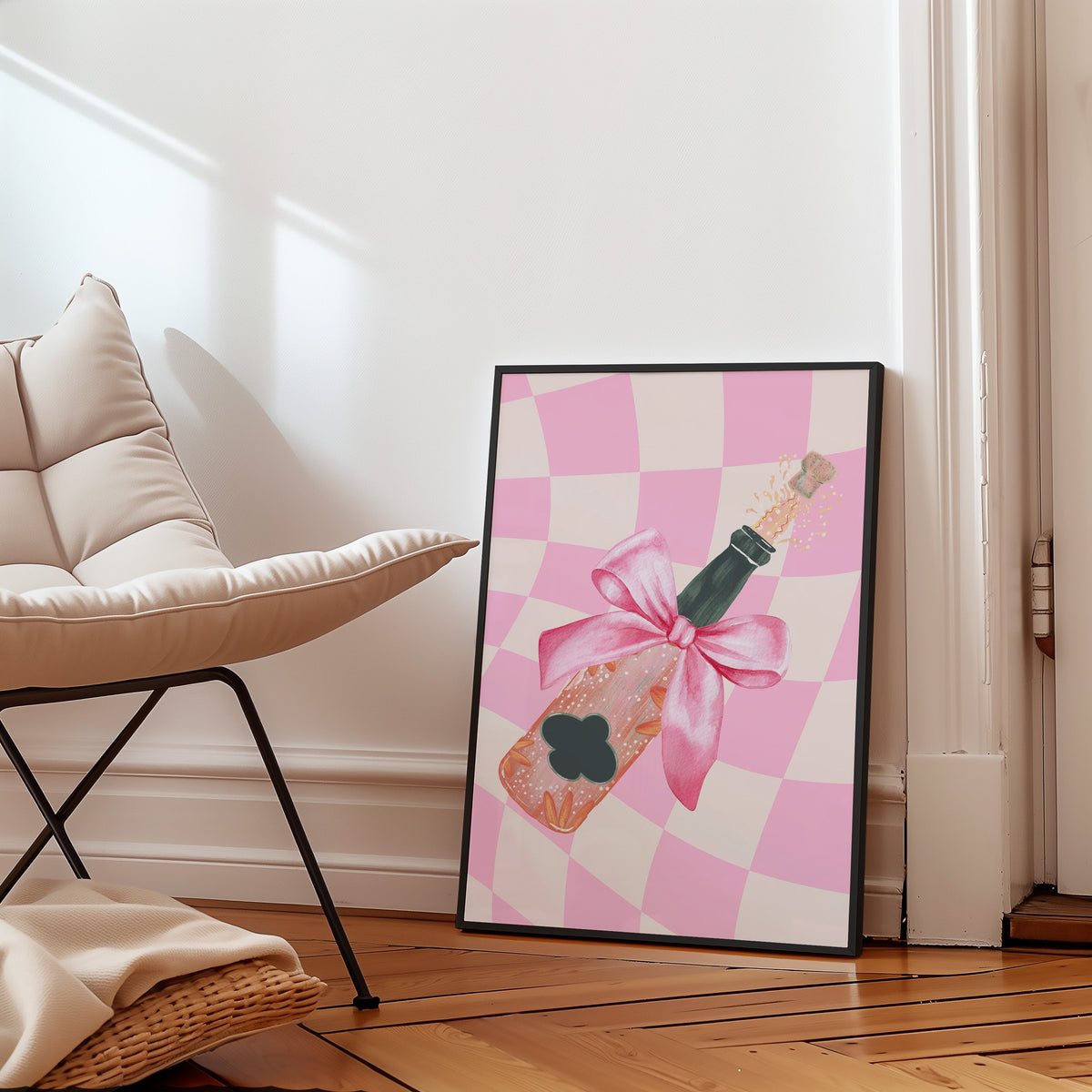 Champagne bottle with a pink bow print in a slim black frame leaning on a herringbone floor beside a cream armchair