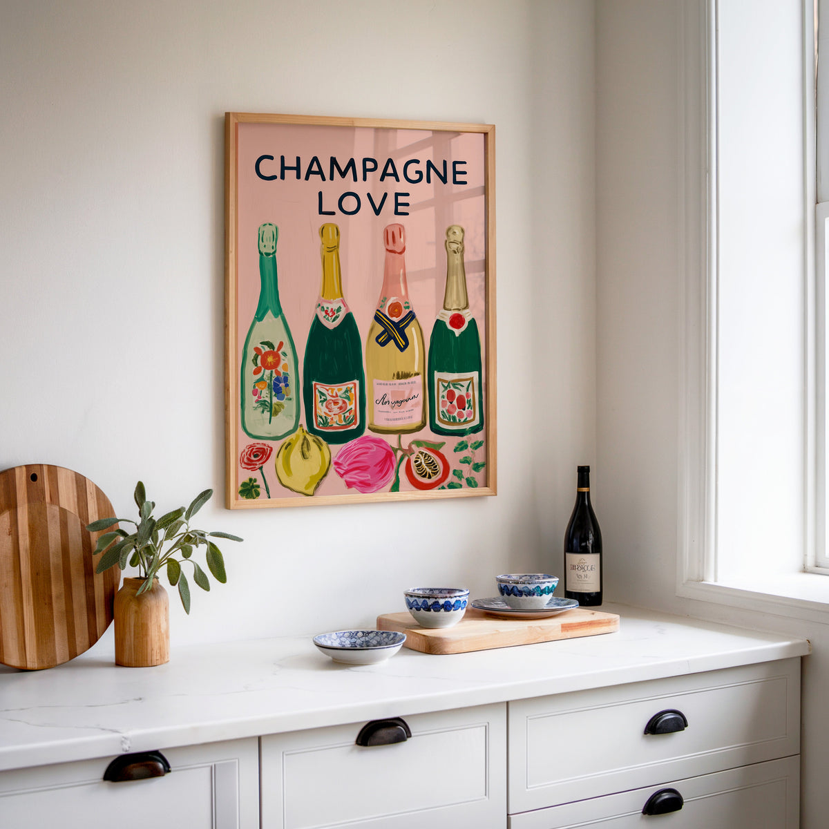 Champagne Love print in an oak frame above a white sideboard with a bottle of wine
