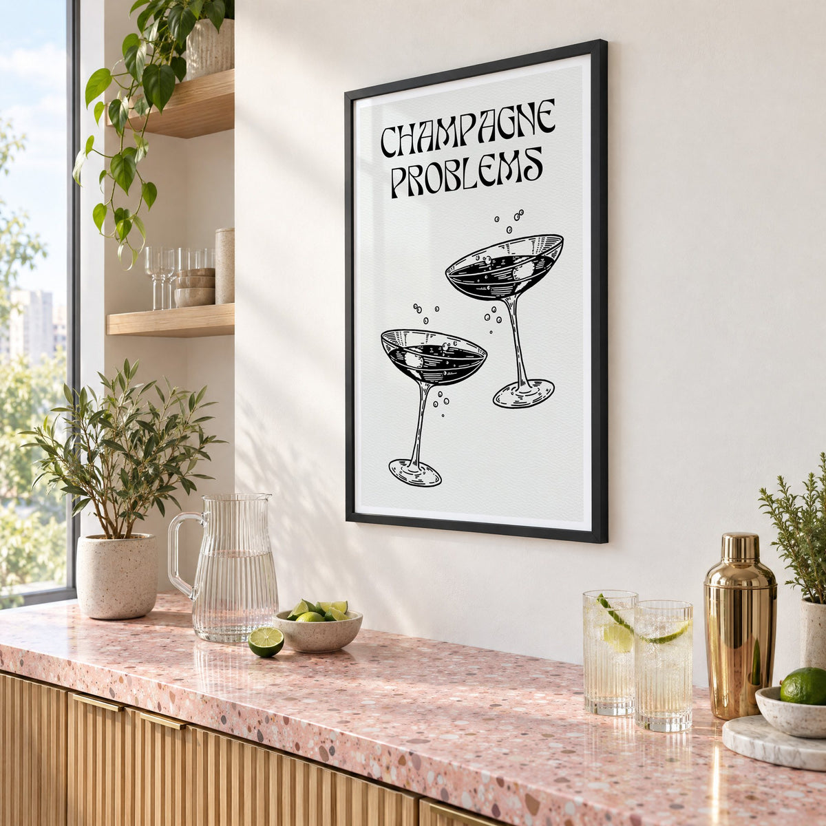 Champagne Problems engraved coupes print in a black frame above a pink terrazzo cocktail counter with limes, seen at an angle