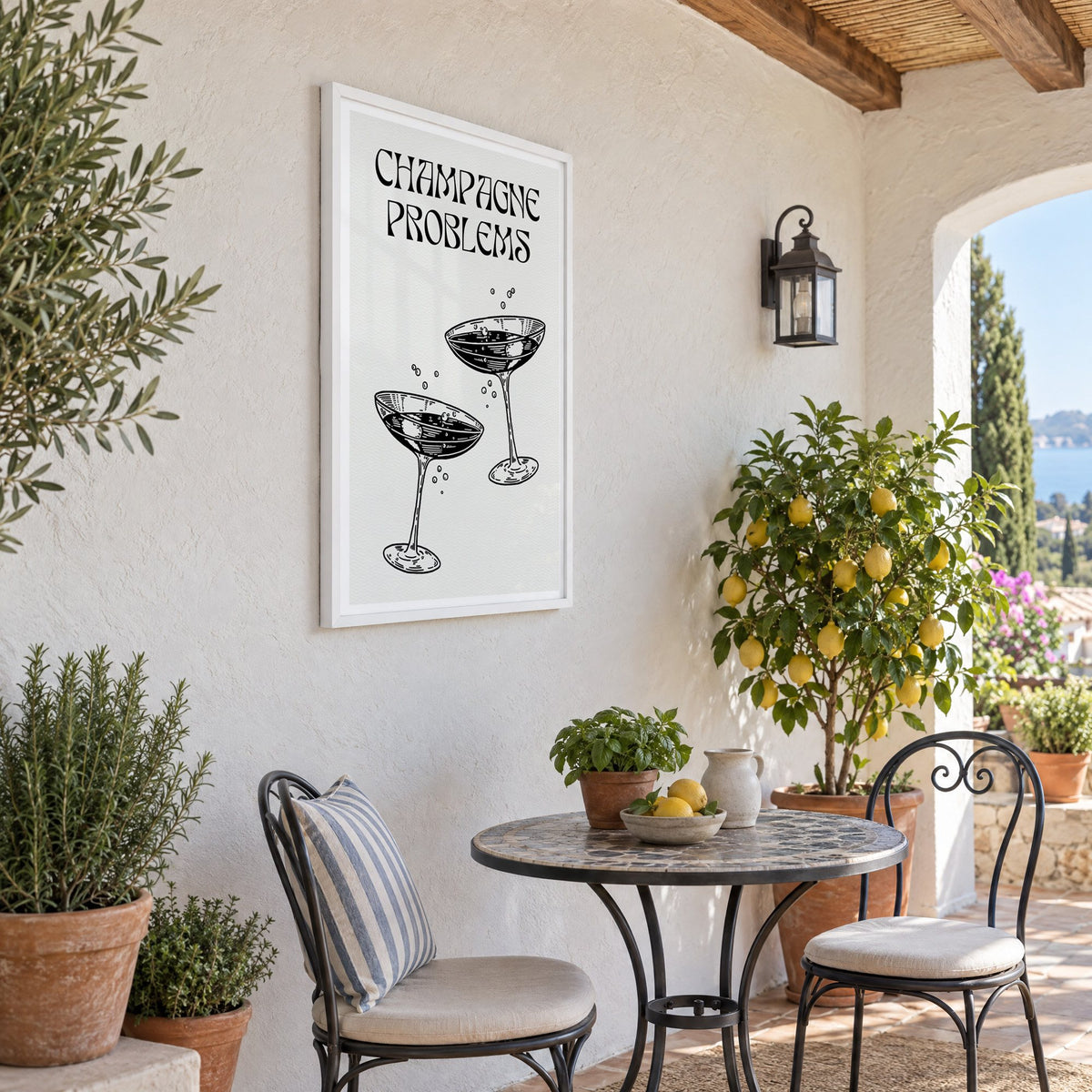 Champagne Problems engraved coupes print in a white frame on a sunny patio wall beside a potted lemon tree