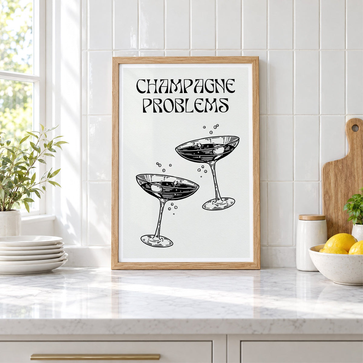 Champagne Problems engraved coupes print in an oak frame leaning on a marble counter by white tiles and lemons