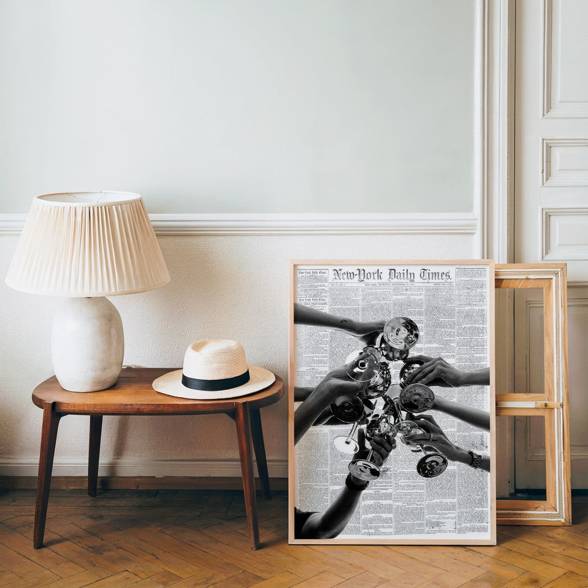 Champagne toast collage on 1851 newspaper print in a pine frame on a wood floor beside a lamp and straw hat