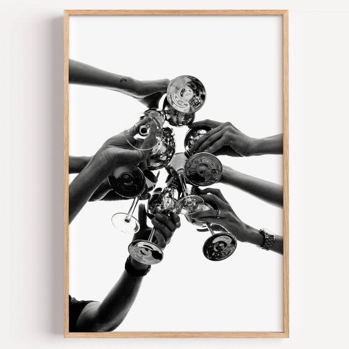 Hands toasting champagne coupes on white print in a light oak frame on a plain white wall