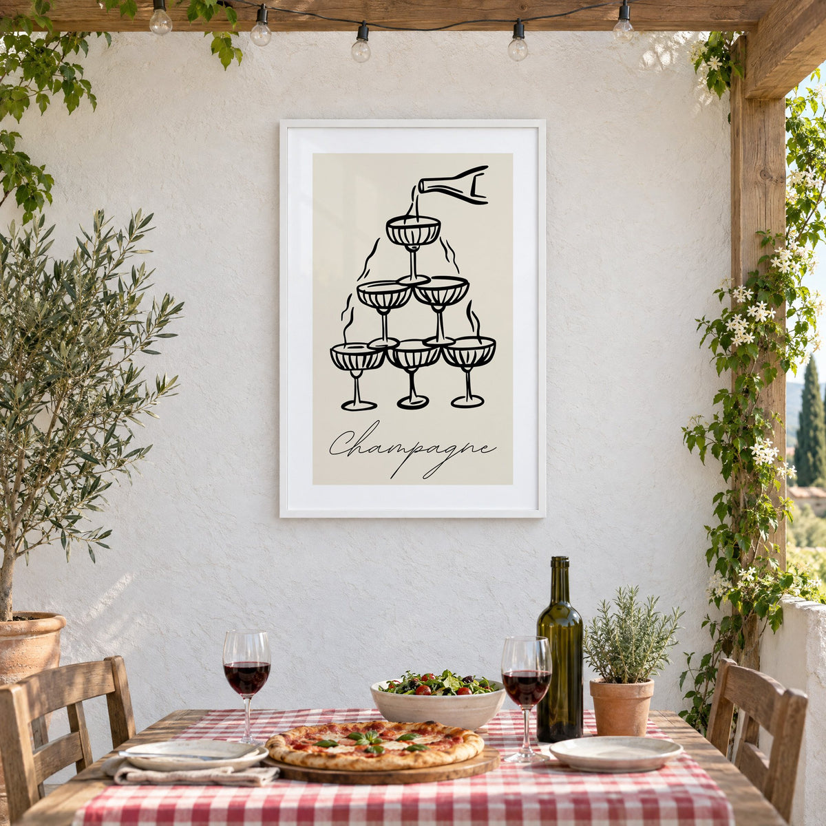 Champagne coupe tower black line-art print in a white frame on a stucco wall above a checked-cloth table with pizza and red wine