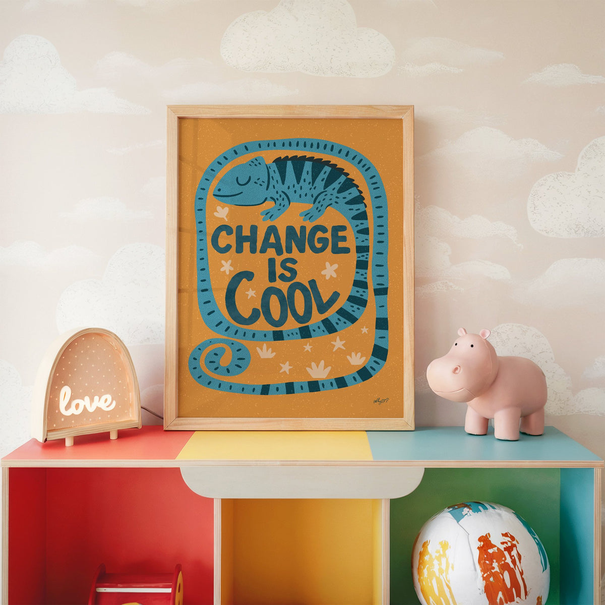 Change is cool chameleon orange nursery print in an oak frame on a rainbow toy shelf against cloud wallpaper