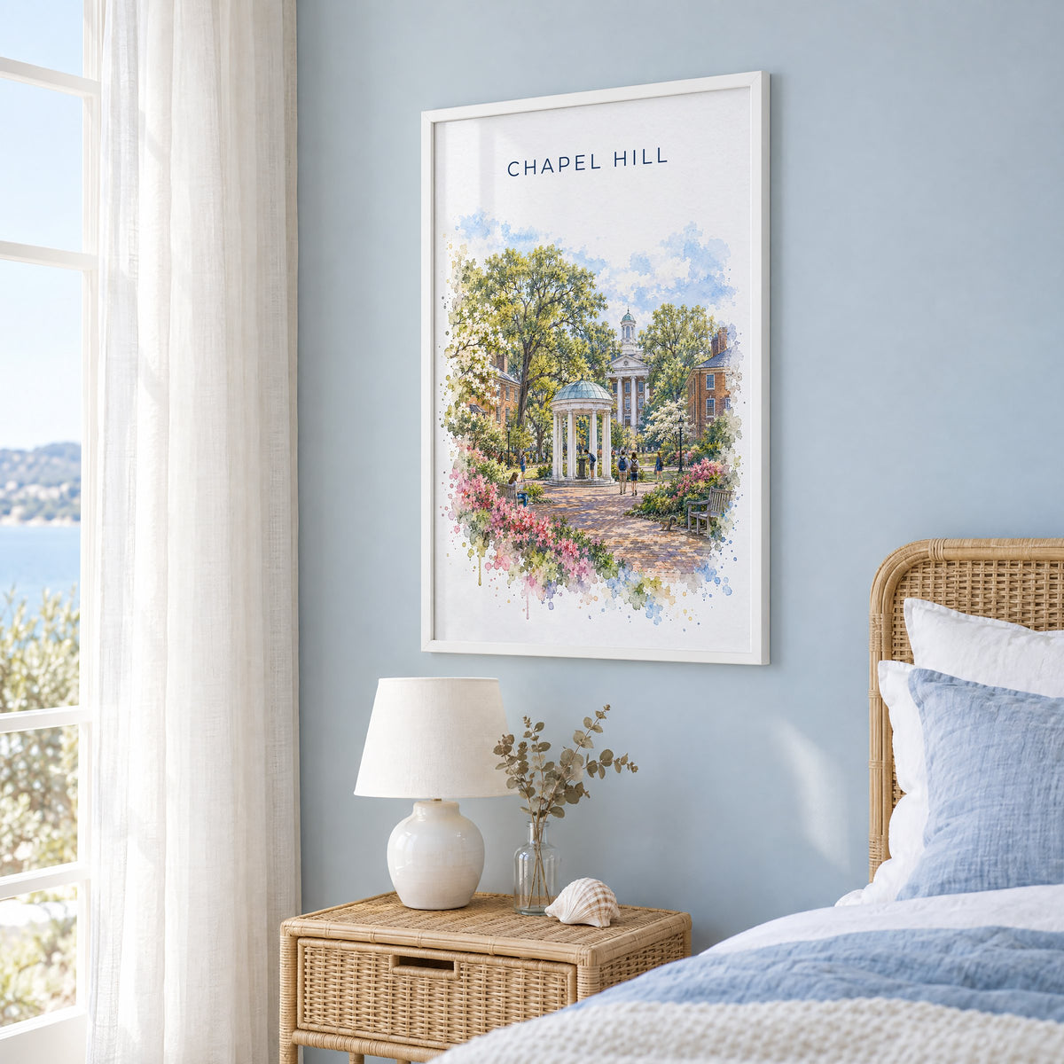 Chapel Hill watercolor print in a white frame on a pale blue wall beside a rattan bed in a coastal bedroom