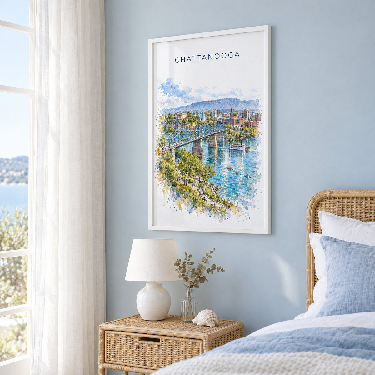 Chattanooga watercolor print in a white frame on a pale blue wall beside a rattan bed in a coastal bedroom