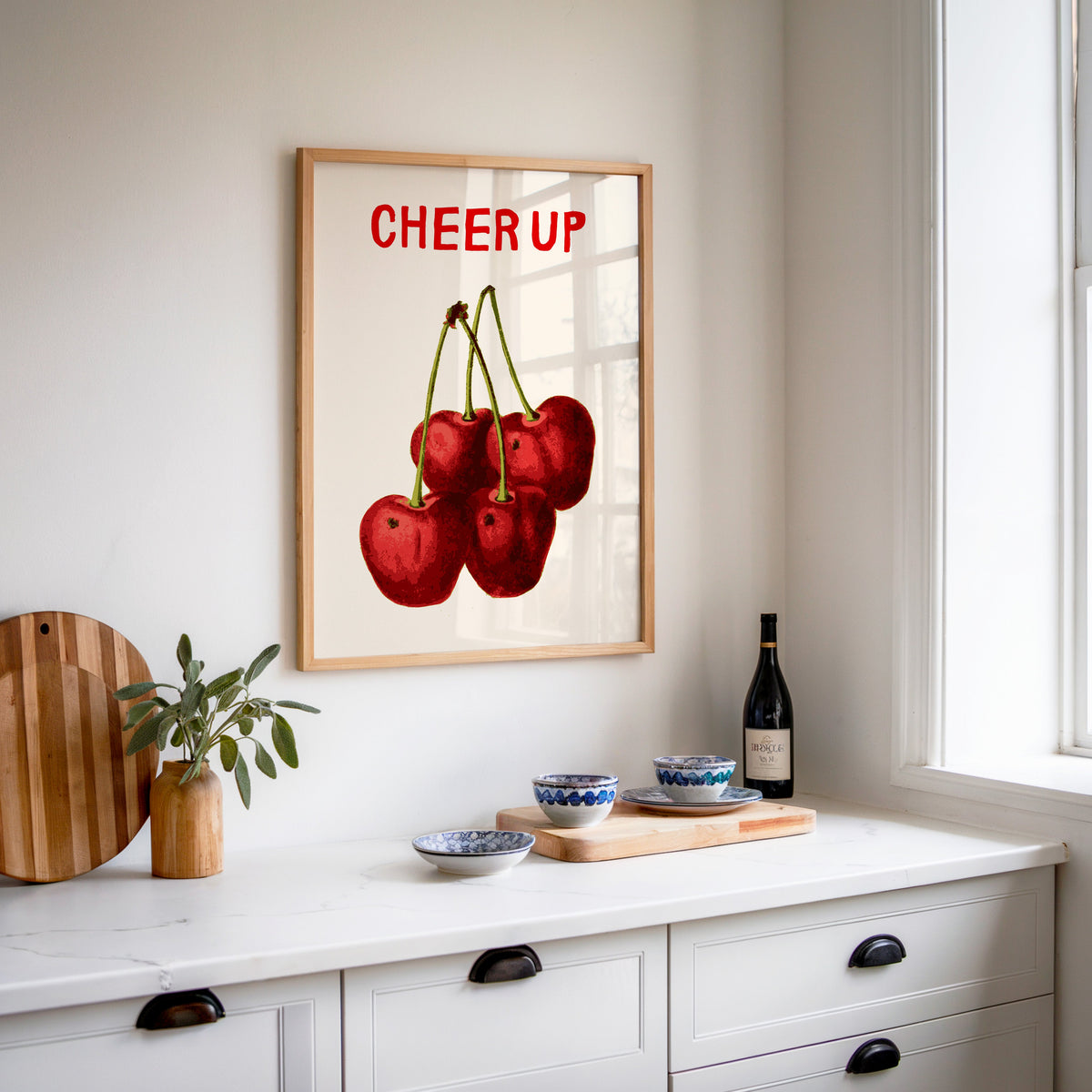 Cheer Up cherries print in an oak frame above a white kitchen sideboard with a wine bottle and wooden board