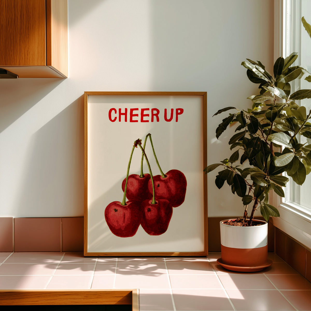 Cheer Up cherries print in a golden oak frame leaning on a pink tiled ledge in strong afternoon sun