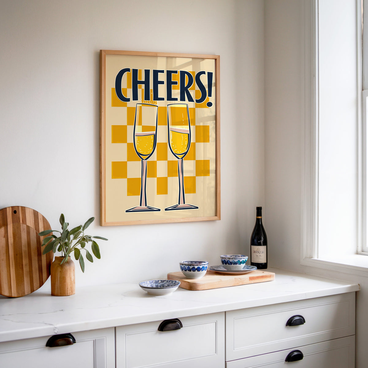 Cheers champagne flutes print in an oak frame above a white sideboard with a bottle of wine
