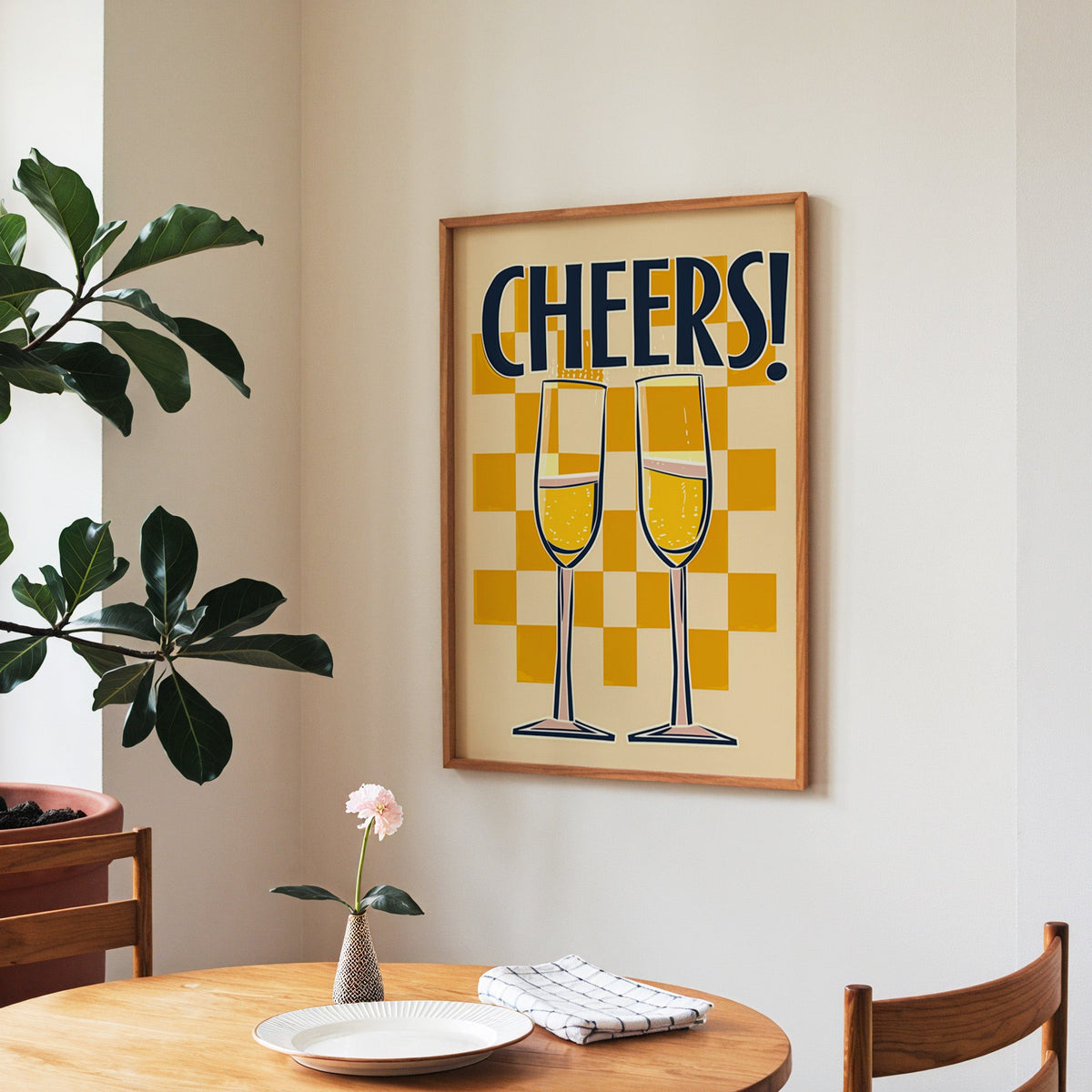 Cheers champagne flutes print in a walnut frame above a round table beside a fiddle-leaf fig