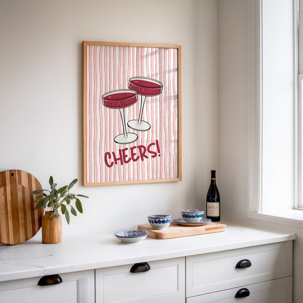 Cheers coupes print in an oak frame above a white sideboard with a bottle of wine