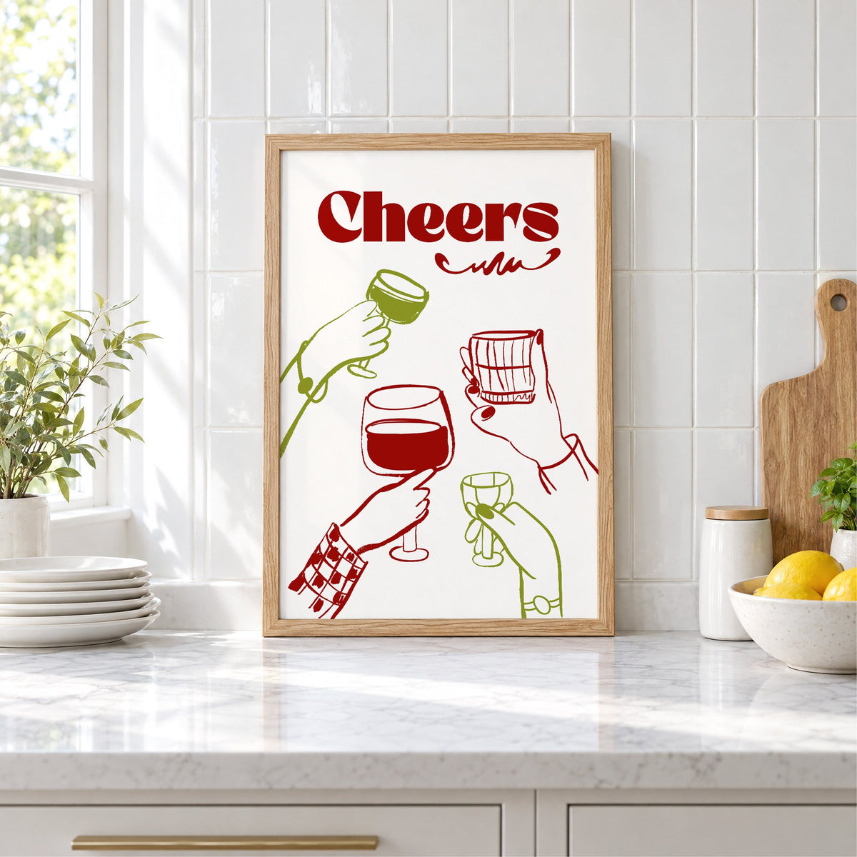 Cheers red and olive toasting hands print in an oak frame leaning on a marble counter by white tiles and lemons