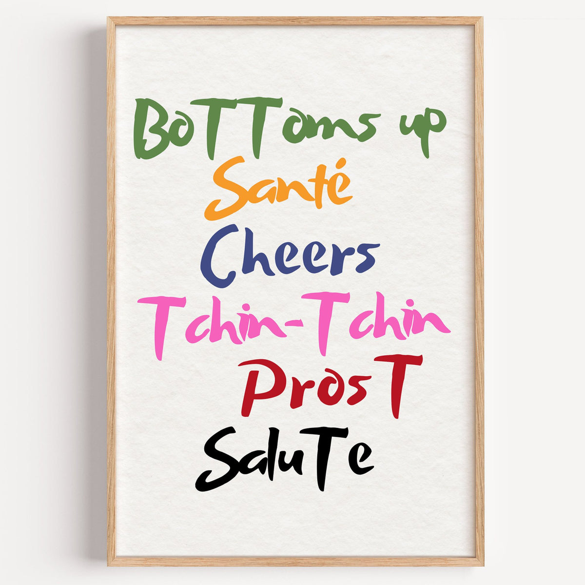 Cheers in six languages brush lettering print in a light oak frame on a plain white wall