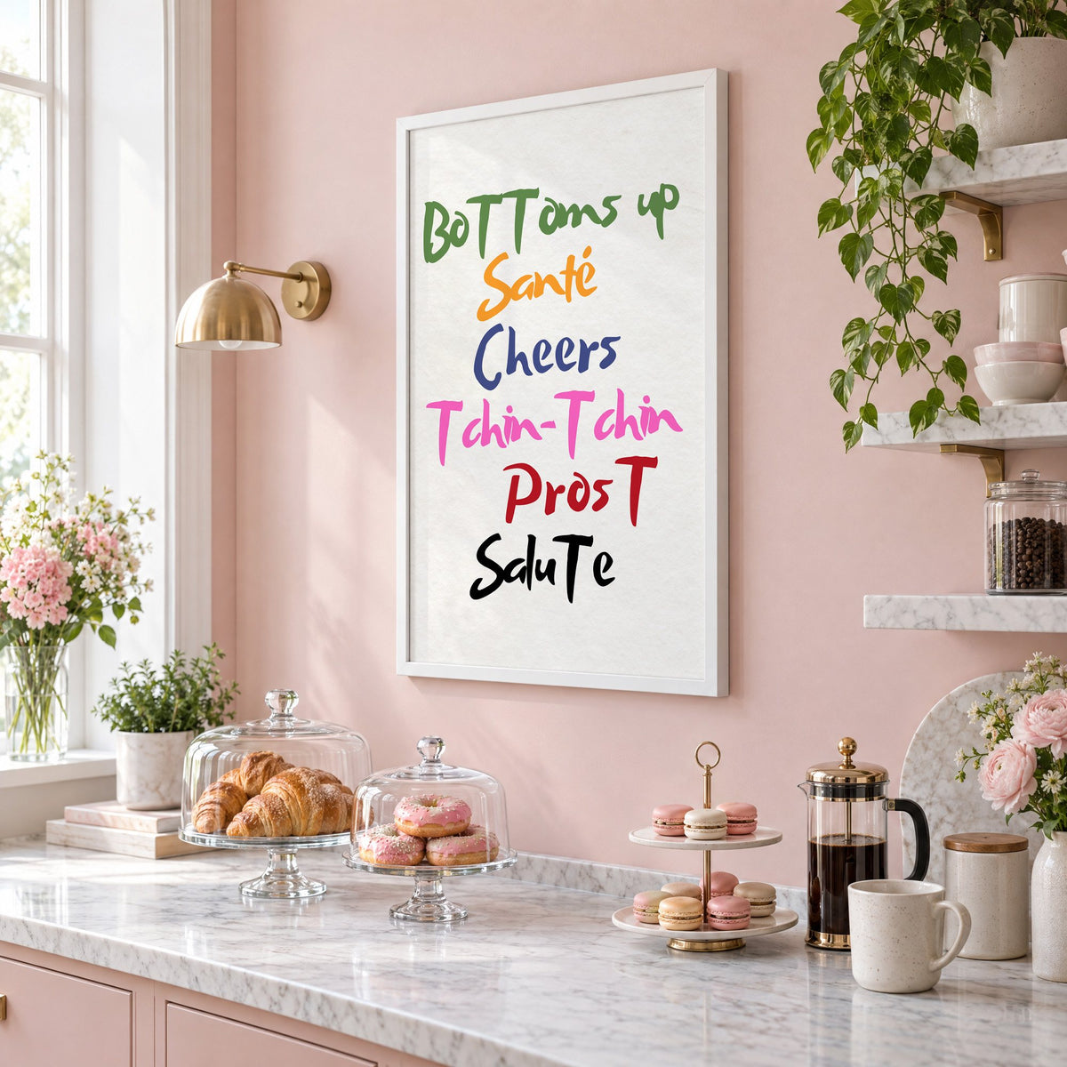 Cheers in six languages brush lettering print in a white frame on a blush pink wall above a patisserie counter with croissants and macarons, seen at an angle