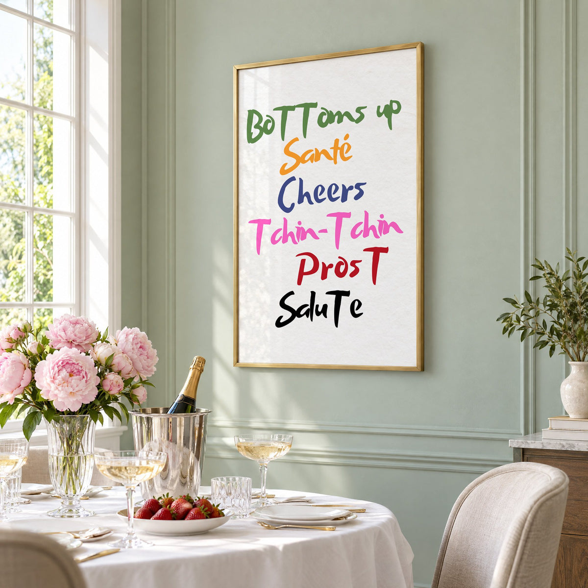 Cheers in six languages brush lettering print in a brass frame on a sage wall above a brunch table with champagne and peonies, seen at an angle