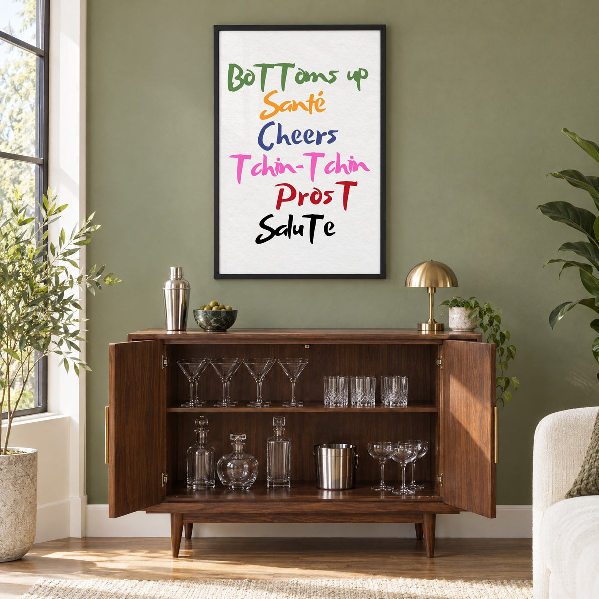 Cheers in six languages brush lettering print in a black frame on an olive green wall above a walnut bar cabinet with glassware