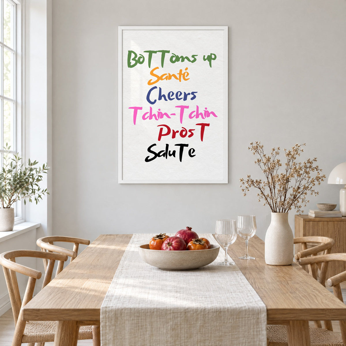 Cheers in six languages brush lettering print in a white frame above a light oak dining table with a linen runner and fruit bowl