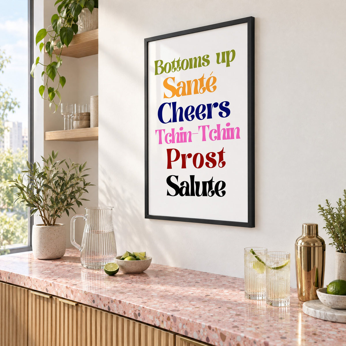 Cheers toasts colorful typography print in a black frame above a pink terrazzo cocktail counter with limes, seen at an angle
