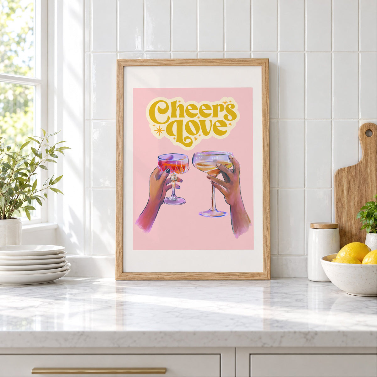 Cheers Love coupe toast print in an oak frame leaning on a marble counter by white tiles and lemons