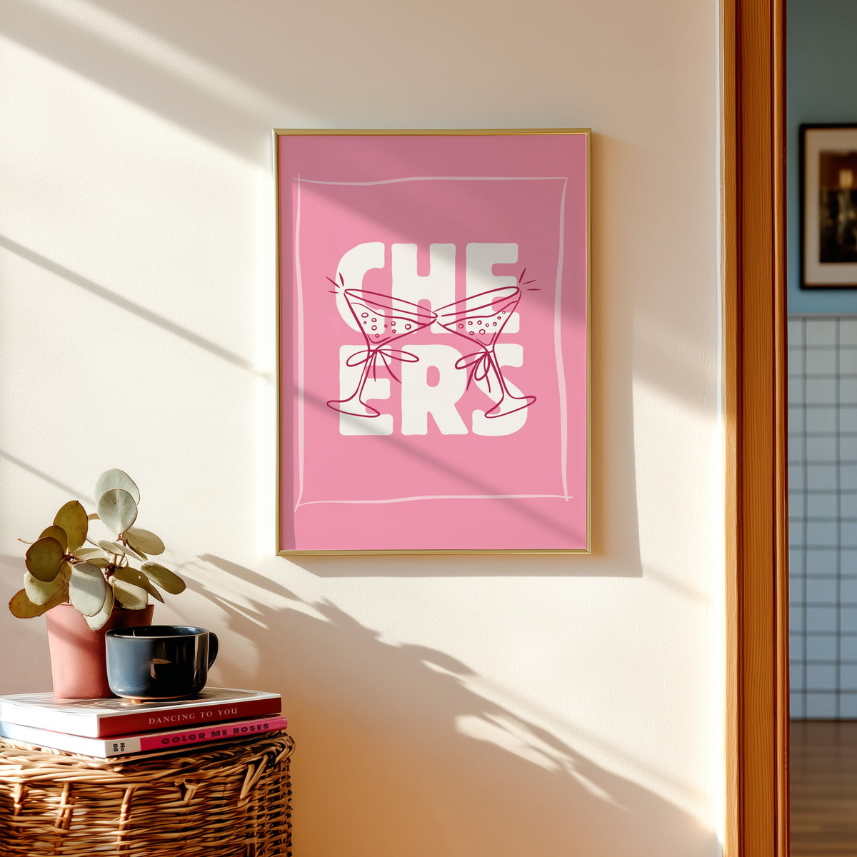 Cheers martini print in a gold frame above a basket of books and a pilea plant