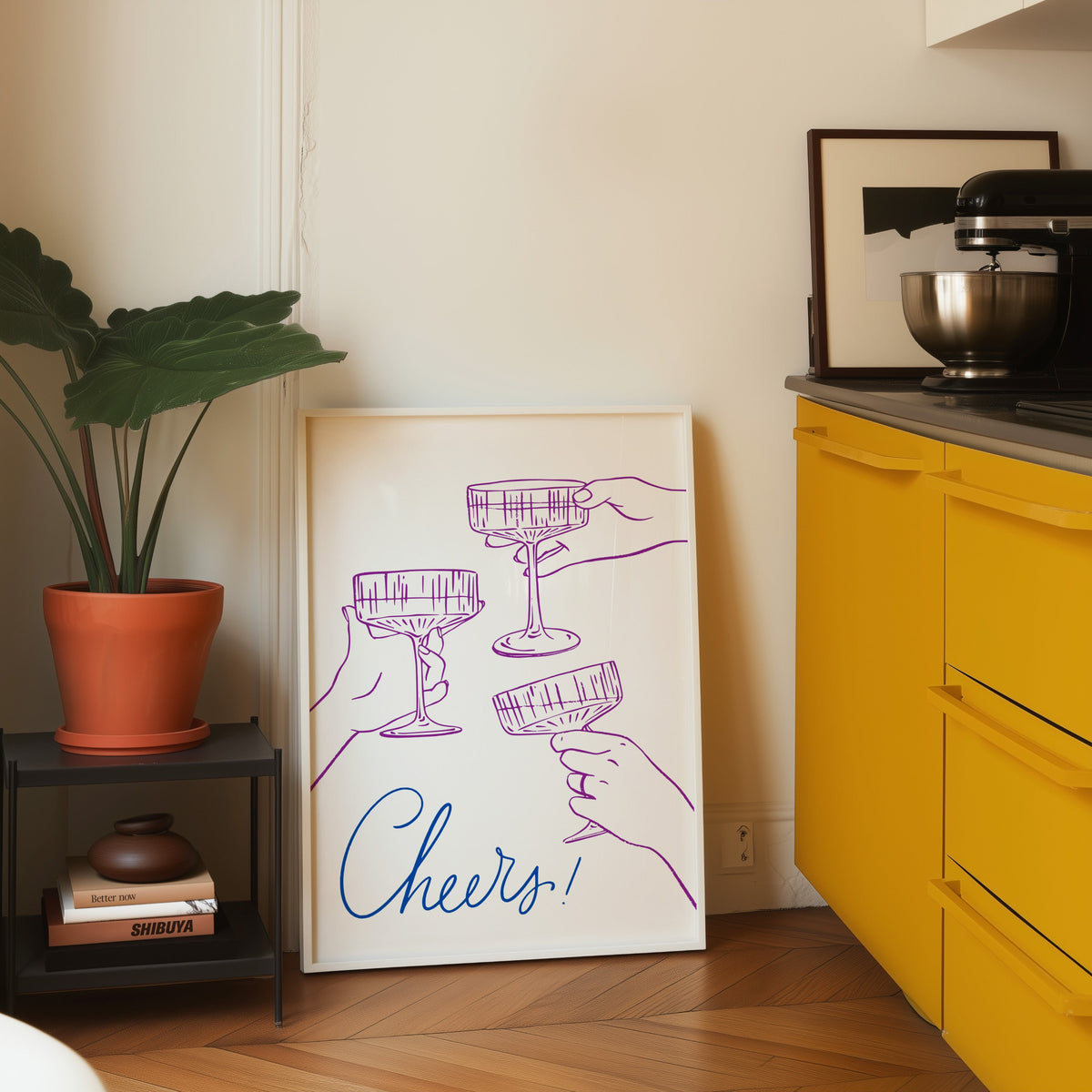 Cheers coupe glasses print in a white frame leaning on parquet beside a bright yellow kitchen