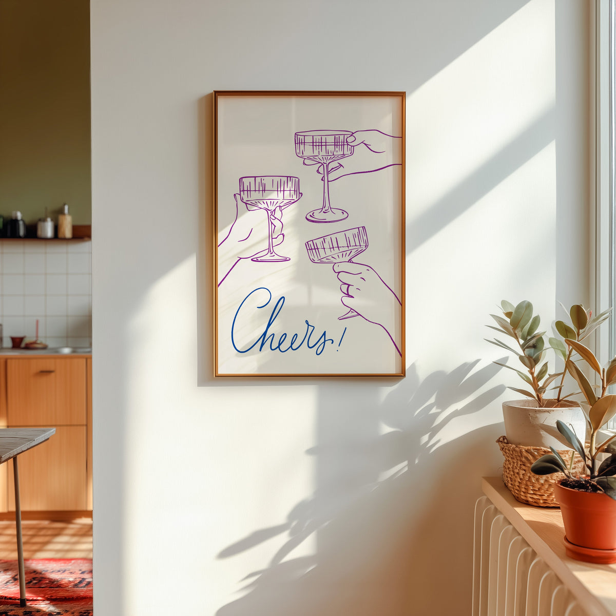 Cheers coupe glasses print shown in a gold frame in sunlight with plants and a kitchen beyond